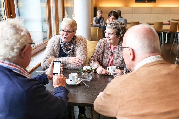 Ältere Personen beim gemeinsamen Kaffeetrinken. Ein Herr trägt ein Hörgerät und hält ein Handy in der Hand.