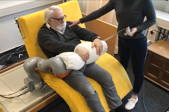 Ein älterer Mann sitzt auf einem Pflegebett, dessen Rückenlehne aufgerichtet ist und das seitlich gedreht wurde. Ein Roboterarm liegt auf seiner Hüfte und eine Person mit Fernbedienung steht neben dem Bett.