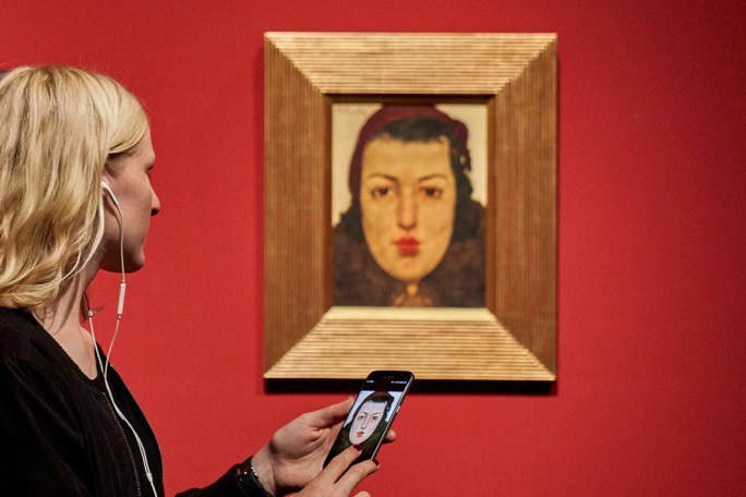 Eine Frau steht mit einem Smartphone in der Hand und Kopfhörern im Ohr vor einem Gemälde.
