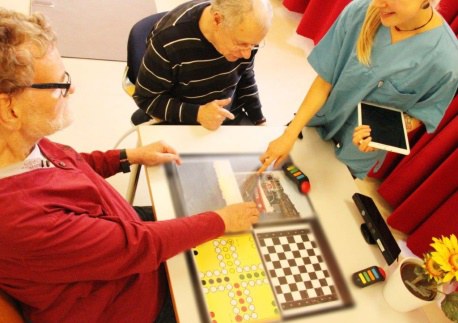 Zwei ältere Personen sitzen am Tisch und eine Ärztin oder Krankenschwester steht mit einem Tablet in der Hand vor ihnen. Auf dem Tisch sind Spiele und Bilder projiziert, auf die eine ältere Dame zeigt.