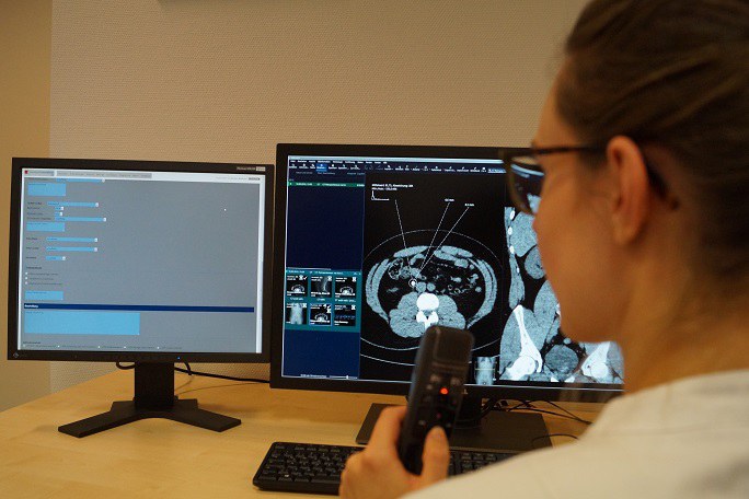 Eine Frau sitzt mit einem Aufnahmegerät vor zwei Computerbildschirmen, auf denen Ergebnisse bildgebender Verfahren zu sehen sind.