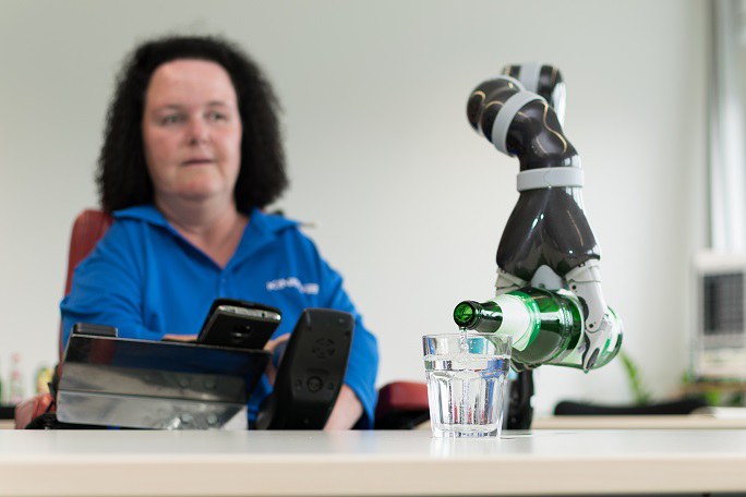 Roboterarm schenkt im Vordergrund Wasser in ein Glas ein. Frau sitzt mit Steuerung im Hintergrund.