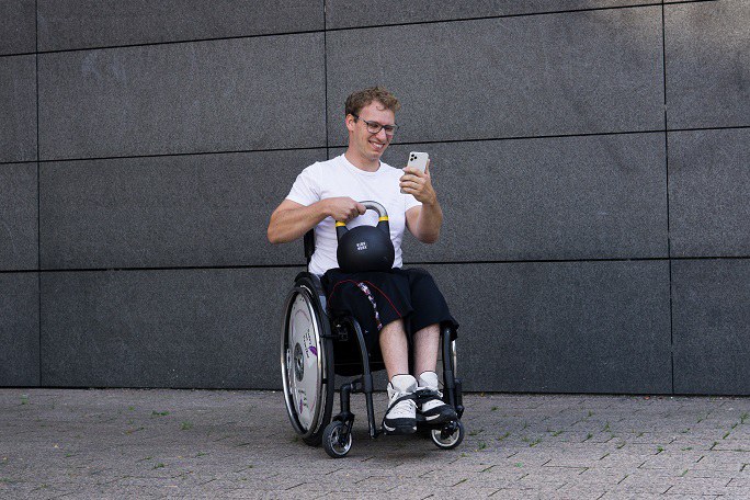 Ein Mann im Rollstuhl hält in der einen Hand eine Kugelhantel und in der anderen ein Smartphone.