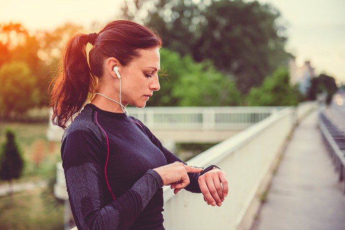 Eine Frau in Sportkleidung und mit Kopfhörern im Ohr steht auf einer Brücke und schaut auf ihre Smart Watch