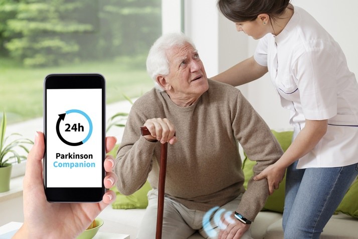 Eine Frau, die einem Man hilft aufzustehen. Im Vordergrund ist ein Handy mit der Aufschrift "Parkinson Companion" zu sehen. 