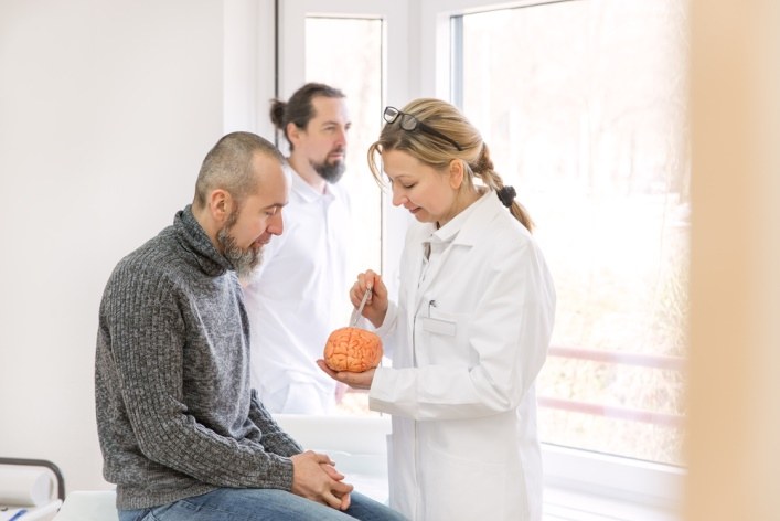 Ein Ärztin spricht mit einem männlichen Patienten. Die Ärztin zeigt mit einem Kugelschreiber auf ein Gehirnmodell.