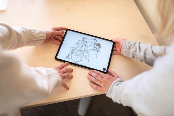 Auf einem Tablet ist eine Konzeptzeichnung einer Person im Rollstuhl zu sehen, welche von einer Pflegenden Person unterstützt wird