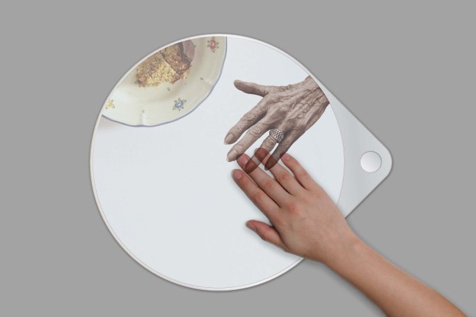 Man sieht eine Hand auf einem Tisch, die eine Projektion einer Hand eines älteren Menschen berührt. In der Projektion ist zudem ein Teller mit Essen zu sehen.