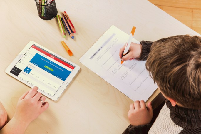 Ein Kind schreibt mit Stift auf Papier und eine zweite Person arbeitet daneben mit einem Tablet