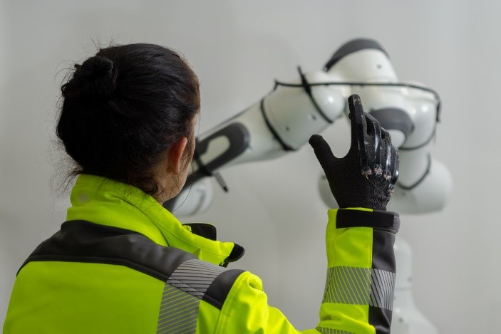 Eine Frau hält einen Arm hoch und man sieht einen taktilen Handschuh auf der Hand. In der Ferne sieht man eine Maschine. 