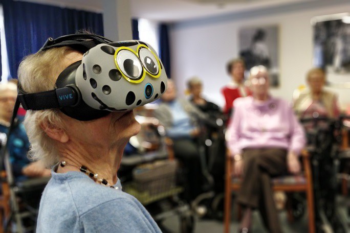 Im Vordergrund ist eine ältere Frau mit einer VR-Brille abgebildet. Im Hintergrund sitzen weitere ältere Menschen.