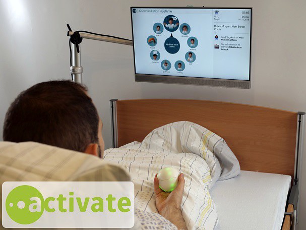Patient in einem Krankenbett vor eine Bildschirm mit einem ballförmigen, leuchtenden Eingabegerät in der Hand
