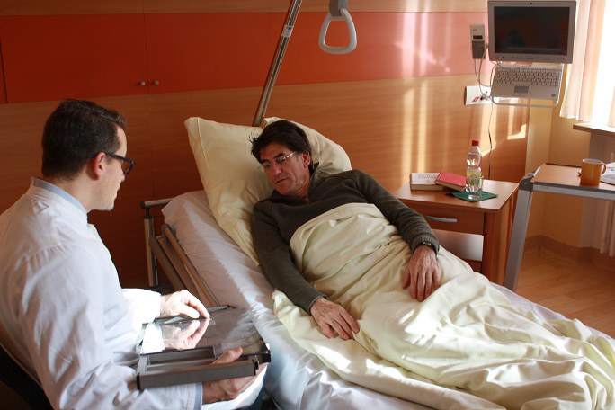 Ein Arzt sitzt mit einem großen, flachen Gerät in der Hand neben dem Krankenbett eines Patienten
